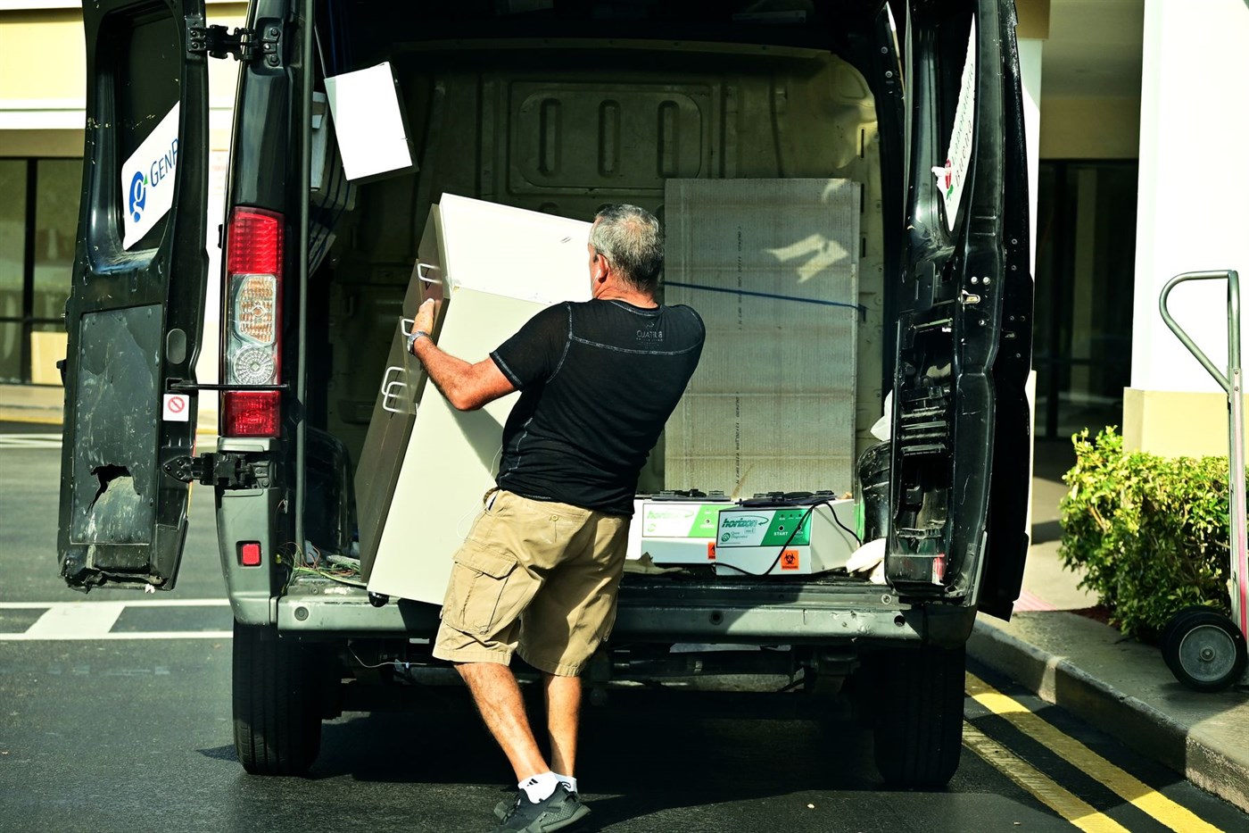 A man lifting a metal cabinet into the back of an open work van in a parking lot