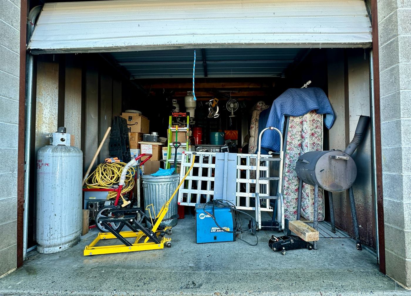 A garage packed to the door with a mattress, ladders, a propane tank, a hand truck and stacked boxes