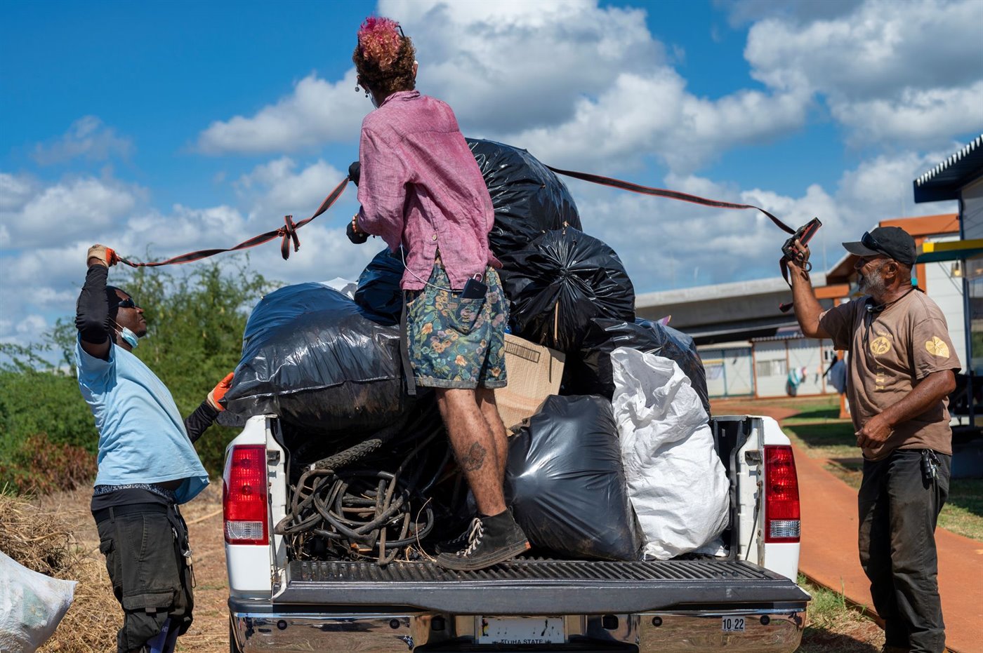 Bagged and loose junk being strapped down in the bed of a pickup truck under a blue sky