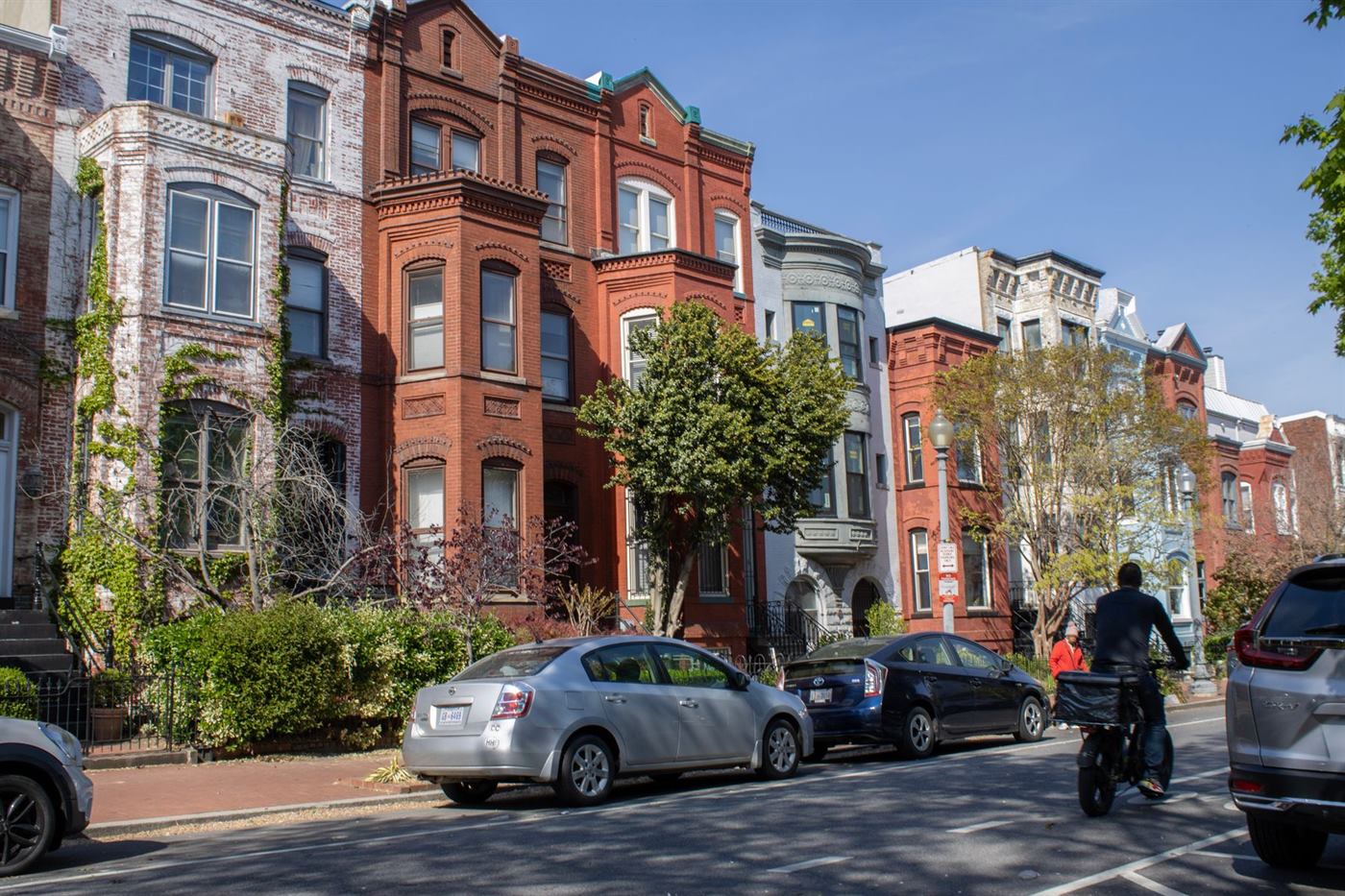 A tree-lined Washington DC street of brick and painted rowhouses with cars parked along the curb
